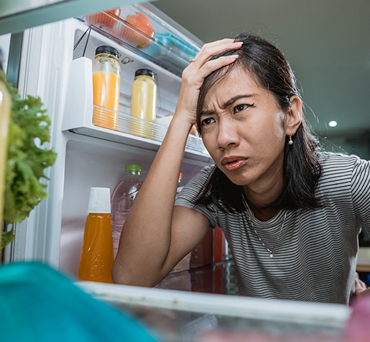 Worried woman looking into an open fridge, noticing food that doesn’t feel cold and suspecting her fridge is failing